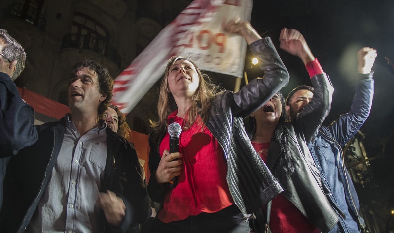 Myriam Bregman, wearing a red shirt and gray blazer, holds a microphone while raising her fist to the sky. Several other people stand behind her in similar poses. They are outside, and it is night.