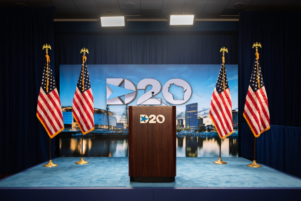 A blue stage with a brown podium in the center, flanked by 2 American flags on each side. Both the podium and the blue backdrop to the stage read D20 in white letters.