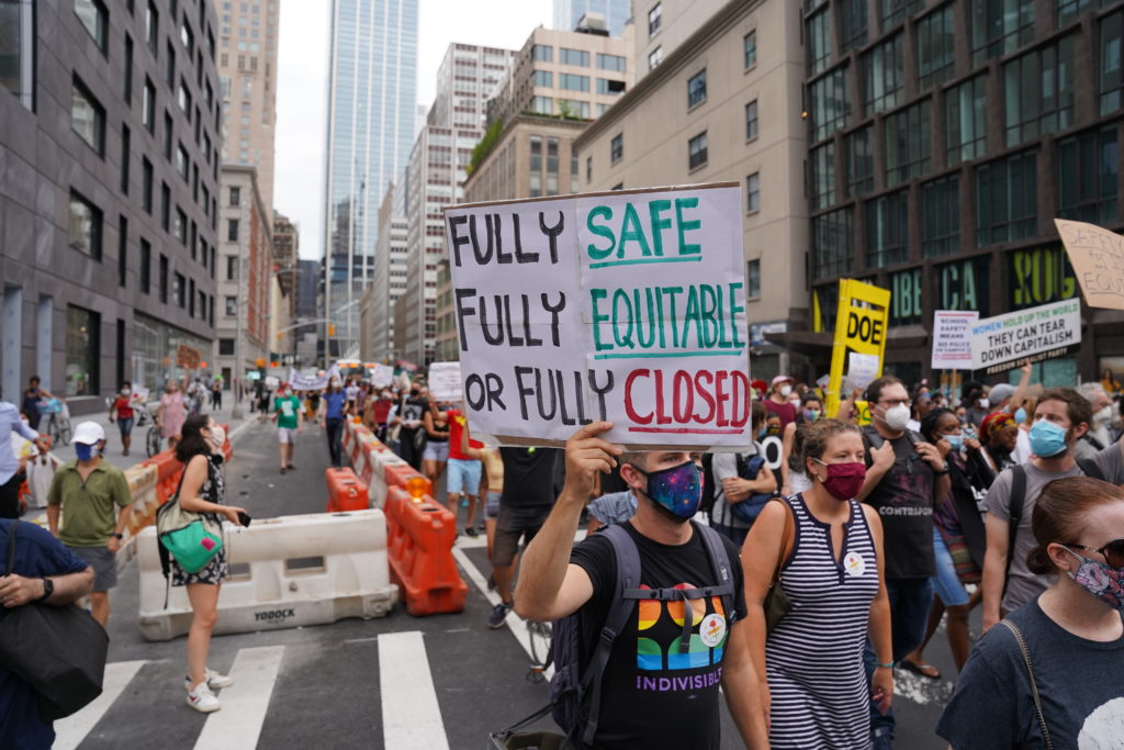 People in a march holding a sign that says "Fully Safe, Fully Equitable, or Fully Closed"
