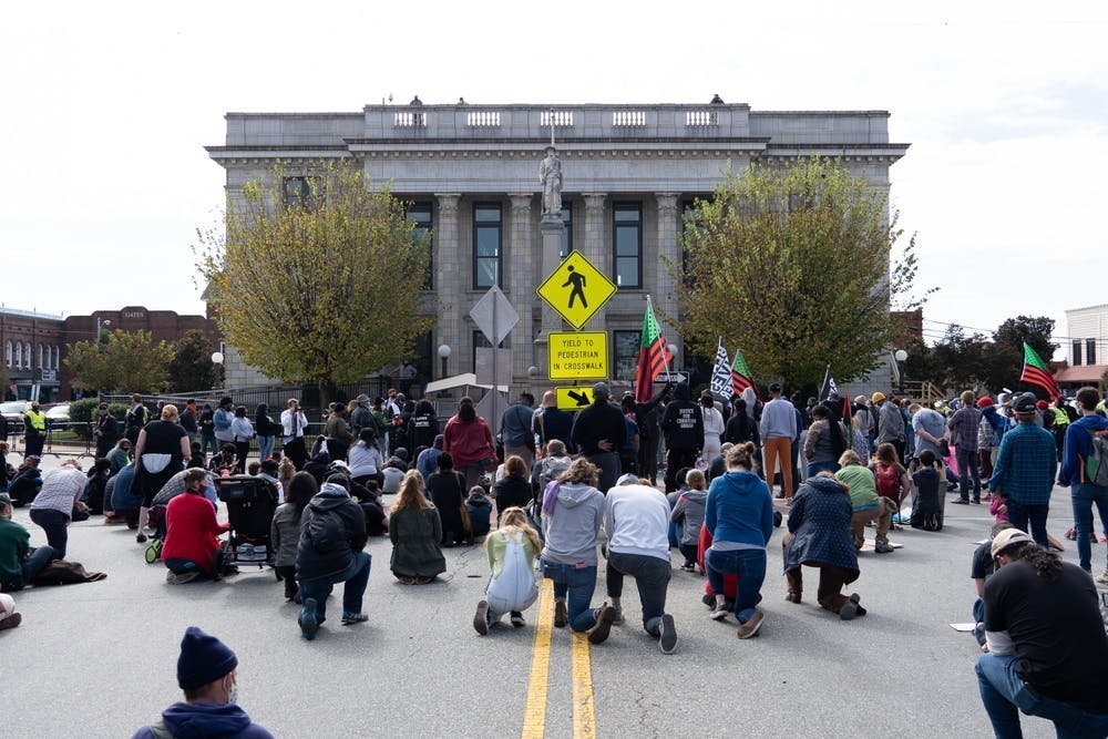 A crowd of semi-socially-distanced people kneel in the middle of a road facing away from the camera. They are facing what appears to be a gray government building with columns.