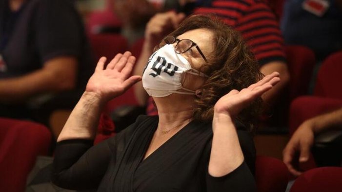 A middle aged woman with curly brown hair that falls to her chin is wearing a white mask, looking upward, and raising her hands, possibly in thanks. Other people (faces not visible) sit behind her on red auditorium-style seats.
