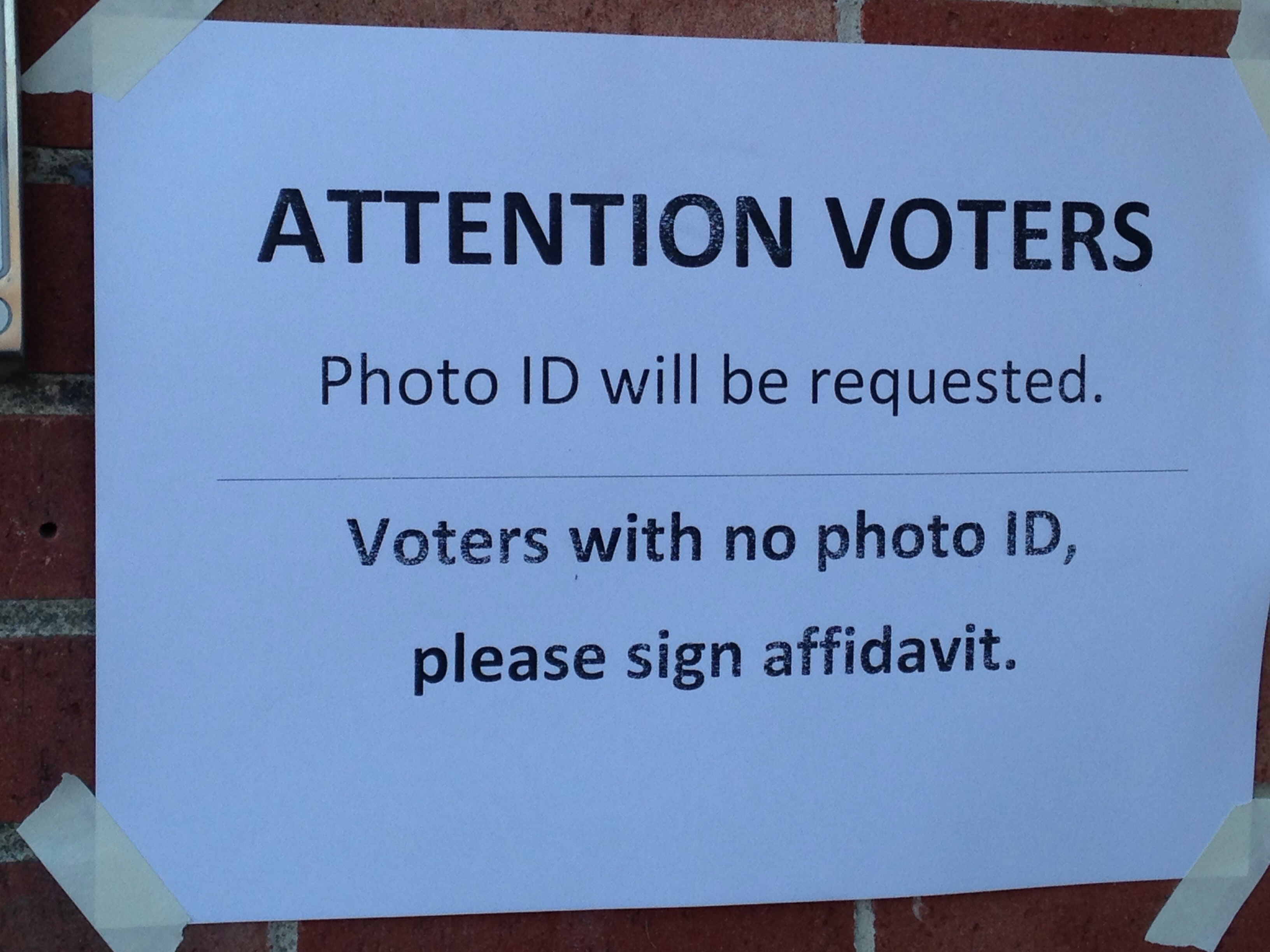 A piece of plain white paper taped to a window reads, "ATTENTION VOTERS: Photo ID will be requested. Voteres with no photo ID, please sign affadavit."