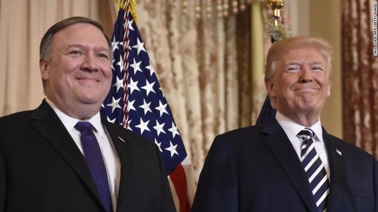 Mike Pompeo and Donald Trump stand beside each other smiling in front of an American flag