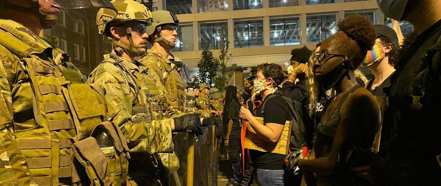 US soldiers stand uniformed wearing helmets facing protesters on the right during the BLM uprisings.