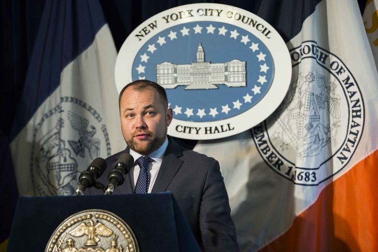 Image of Corey Booker, a white man with a beard in a black suit with a blue tie, in front of a sign that says New York City Council on top and City Hall on the bottom.