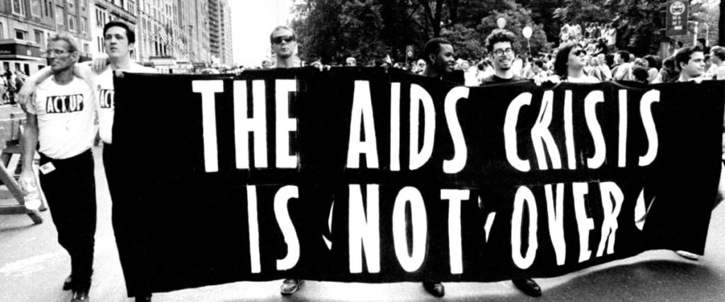 Black and white photo of people marching with a banner that says "The AIDS Crisis Is Not Over"