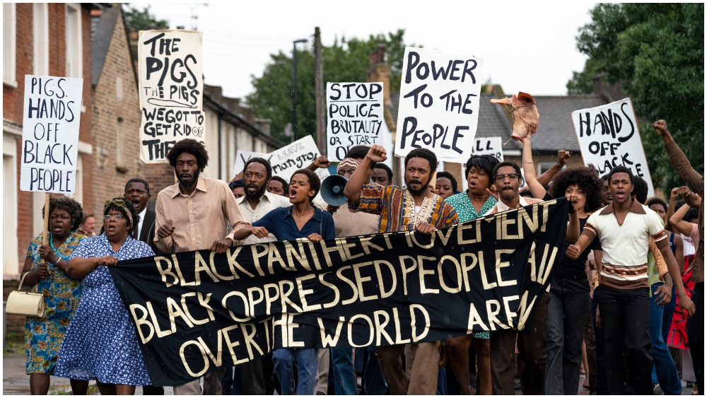 A screenshot from the movie Mangrove. A group of around 20 Black activists march down the street on a bright but cloudy day, carrying signs in protest of police brutality. The banner at the front of the group is black with white text reading " Black Panther Movement (line break) Black Oppressed People (word unreadable due to fold in the banner) (line break) Over the World (word unreadable due to fold in the banner)