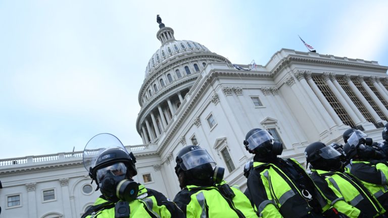 Police in neon yellow vests and shielded helmets stand outside Capitol building.