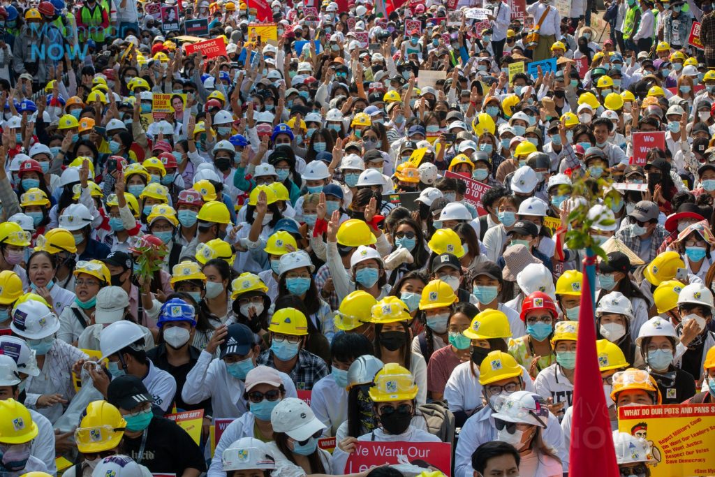 The entire frame of the photo is packed with Myanmarese workers, many of whom are wearing yellow or white hard hats. Most of them are wearing masks, and many are holding one hand in the air, palm forward, with the thumb tucked across the palm.