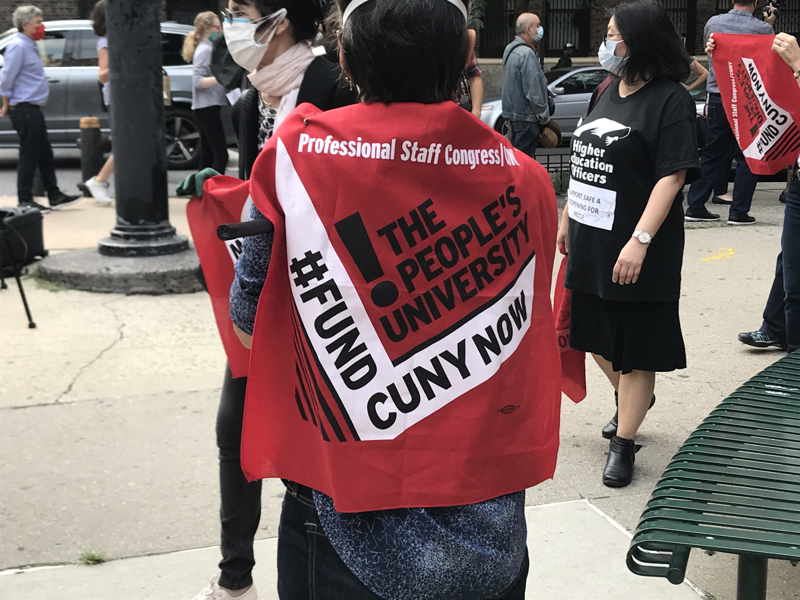 Someone with short dark hair stands with their back to the viewer. On their back, they are wearing a piece of red cloth that says "Professional Staff Congress. The People's University. #FundCUNYNow!" In the background, there are other people marching and carrying similar signs.