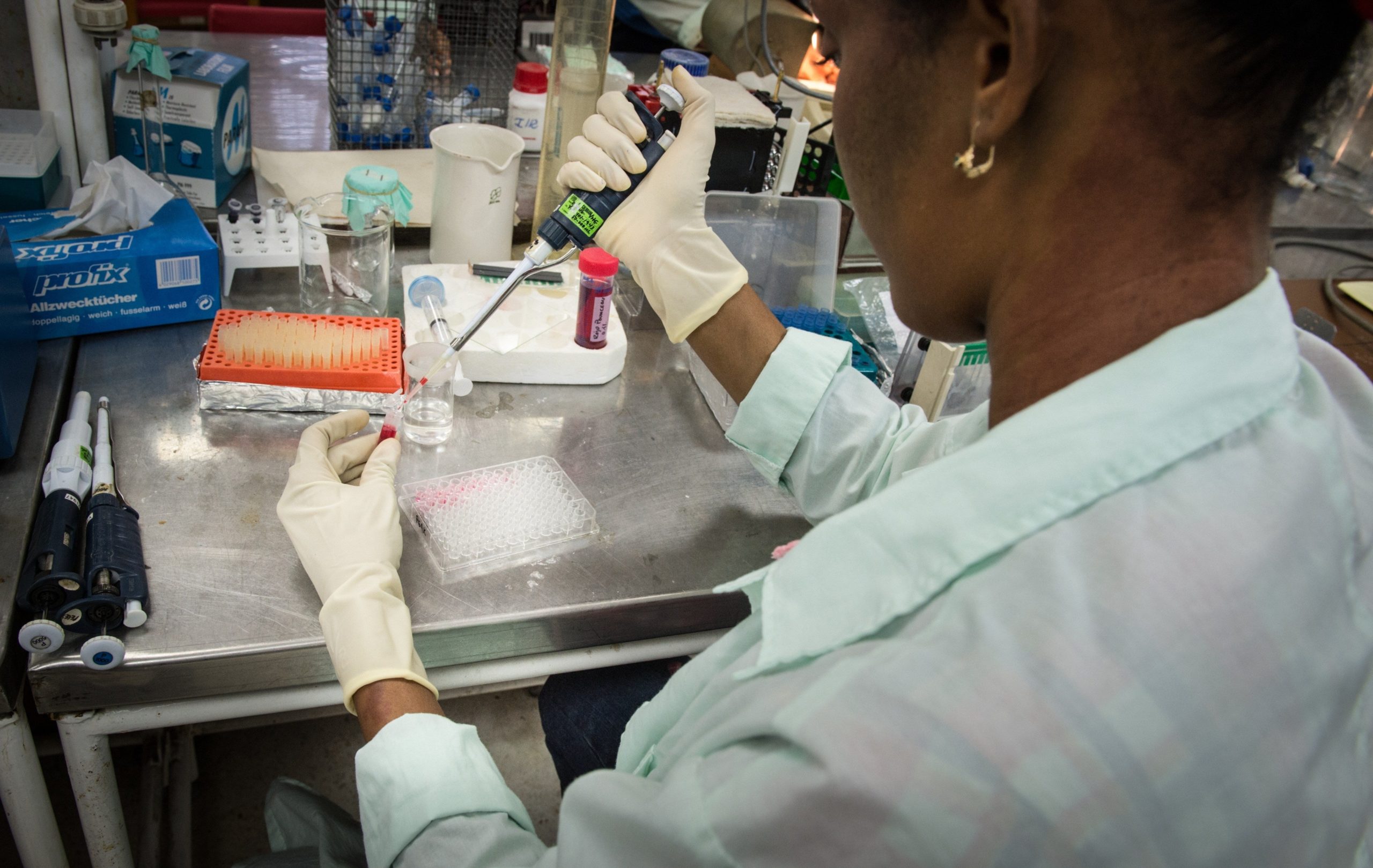 A brown-skinned woman wearing a white lab coat and white gloves is sitting at a laboratory table with a syringe in one hand and a small container in the other