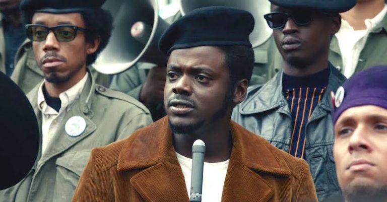 The actor Daniel Kaluuya in character as Fred Hampton. He is standing in front of a microphone looking serious and concerned, with other Black activists standing behind him.