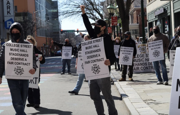 A group of people, mostly white men, mostly wearing dark jackets, jeans, and masks, stand in the street carrying signs that read "College Street Music Hall Stagehands Deserve a Fair Contract." One man is holding his fist into the air.