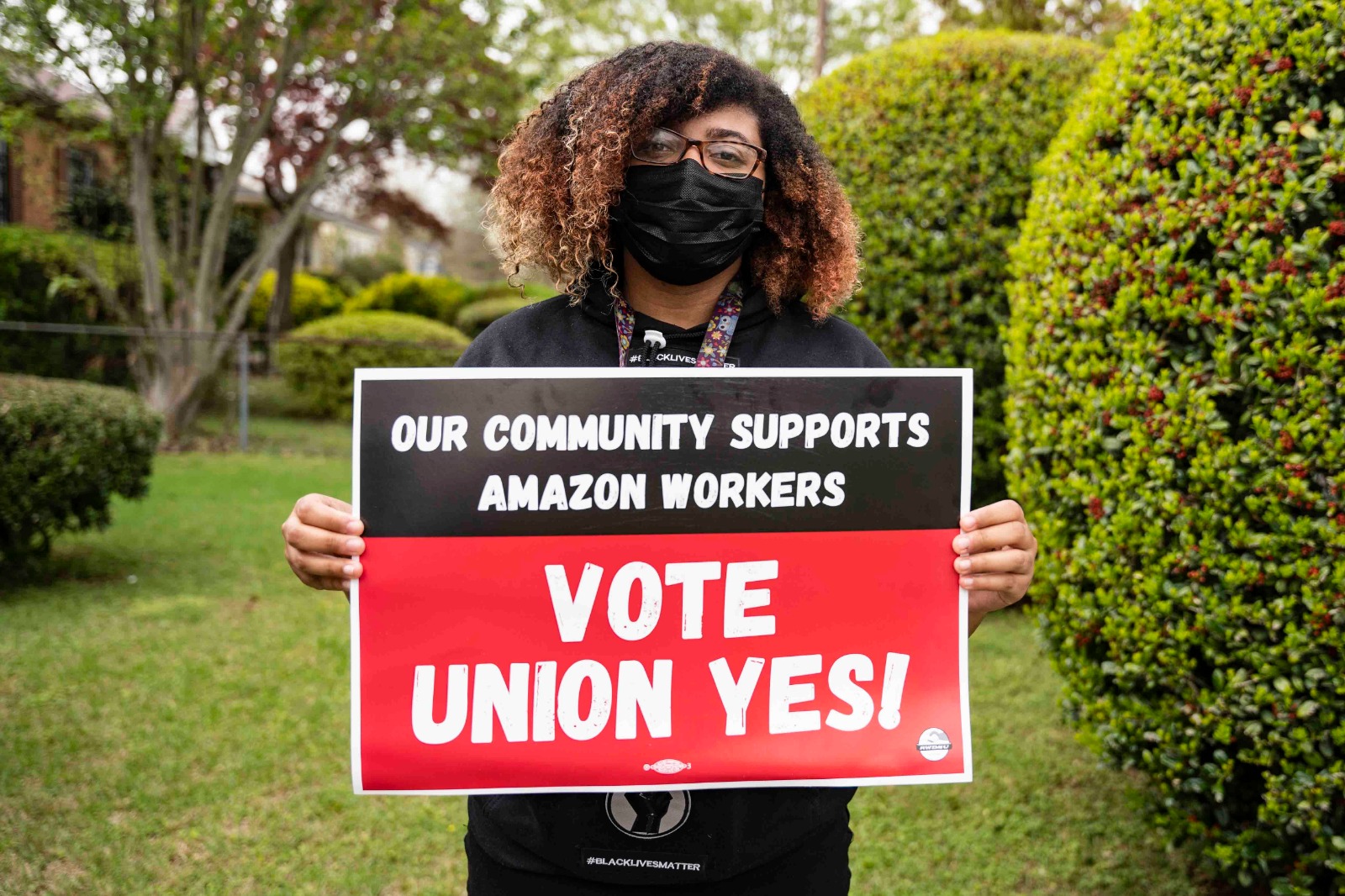 Frances Wallace, a Black woman wearing glasses and a plan black mask, holds a sign that says "Our Community Supports Amazon Workers: Vote Union Yes!"
