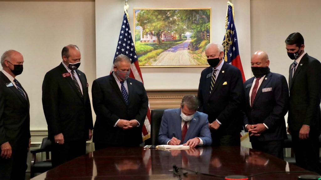 Georgia Governor Brian Kemp sits flanked by 6 white governors while he signs a voter suppression bill into law. In the background is a painting that depicts a slave plantation.