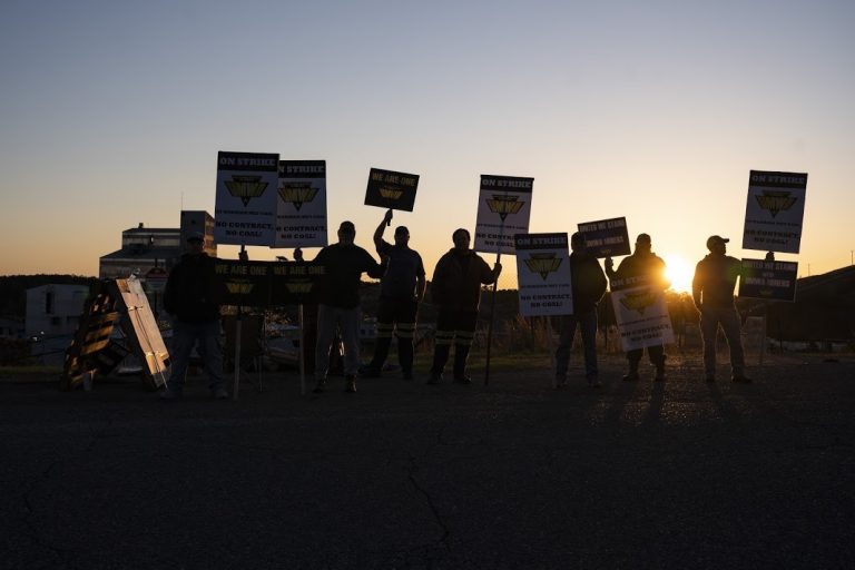 With the sun setting in the background, striking coal miners with Warrior Met stand holding up signs. They look silhouetted.