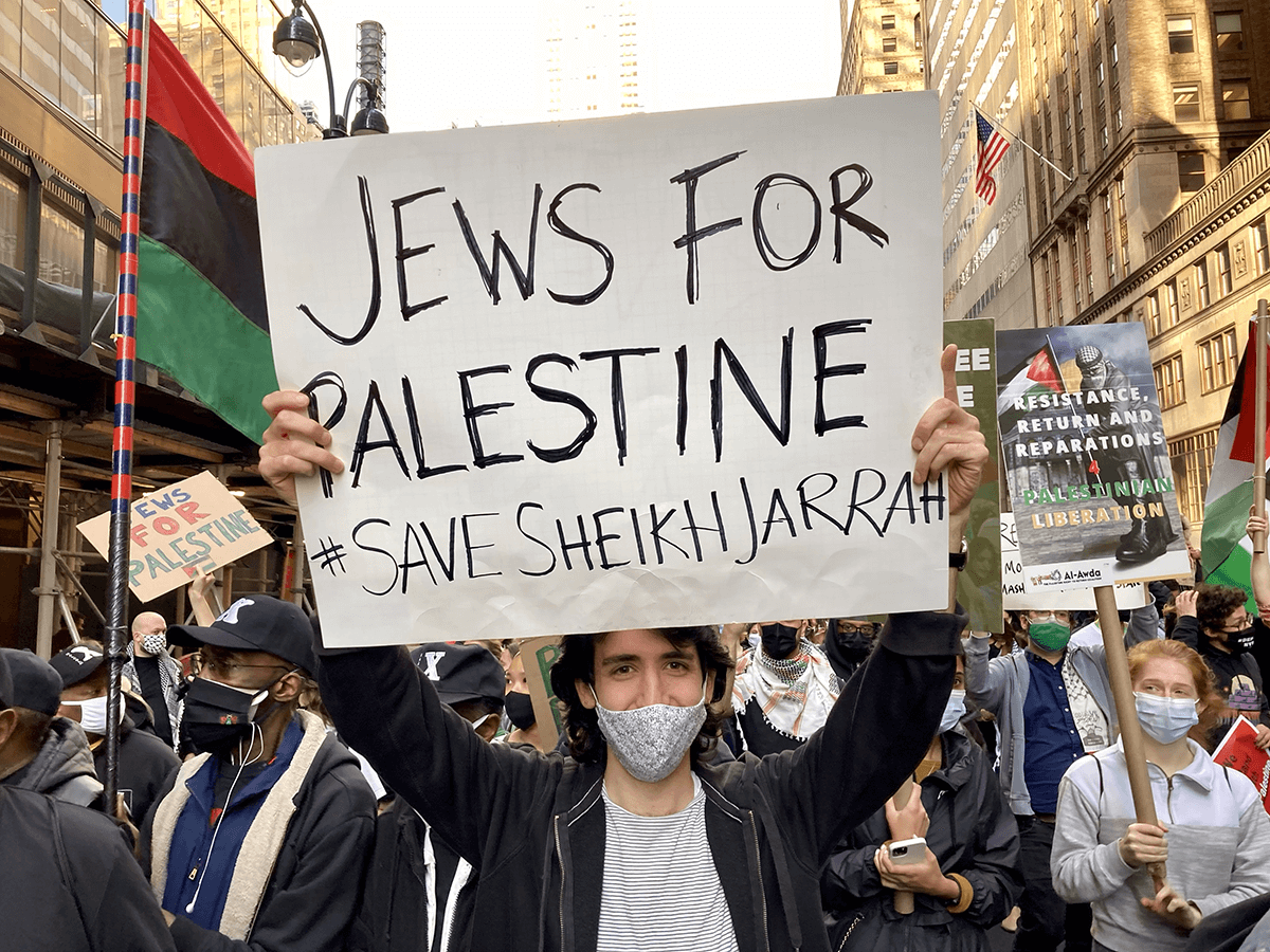 A young adult wearing a medical mask holds a sign above their head that reads "JEWS FOR PALESTINE, SAVE SHEIKH JARRAH"