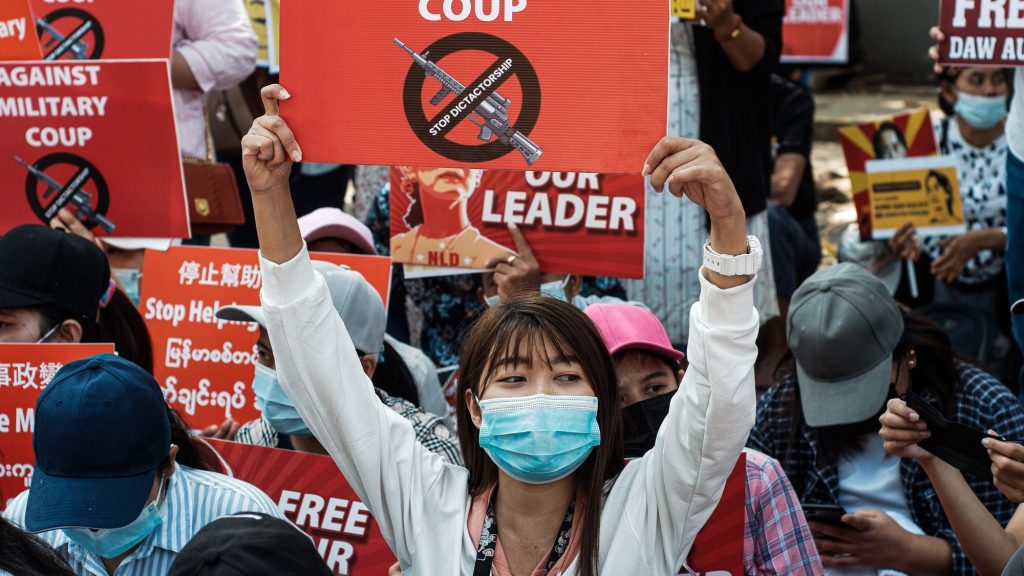 Person wearing a blue face mask holds a red sign that says "stop dictatorship" in front of a crowd of protesters.