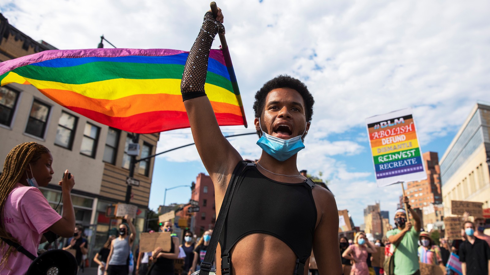 A Black person in a black crop top is holding a rainbow flag. They are in front of a parade.
