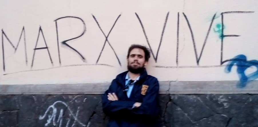 Cuban Marxist Frank García Hernández stands with his arms crossed against a wall that reads "Marx Vive" (Marx Lives)