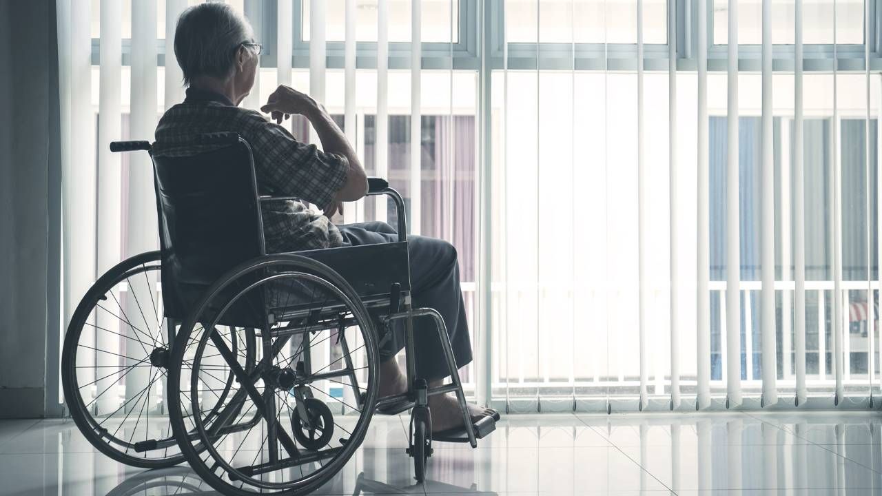Elderly person sitting in a wheelchair with their back facing the camera in front of a window with the shutters open.