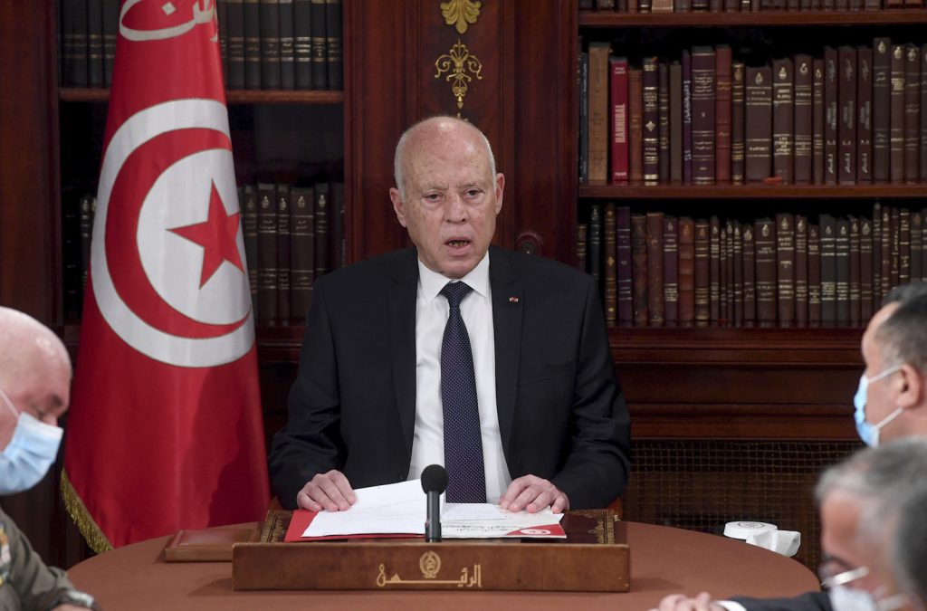 Tunisian President Kais Saied sits at a conference table wearing a suit. The Tunisian flag and a wall of books are in the background.