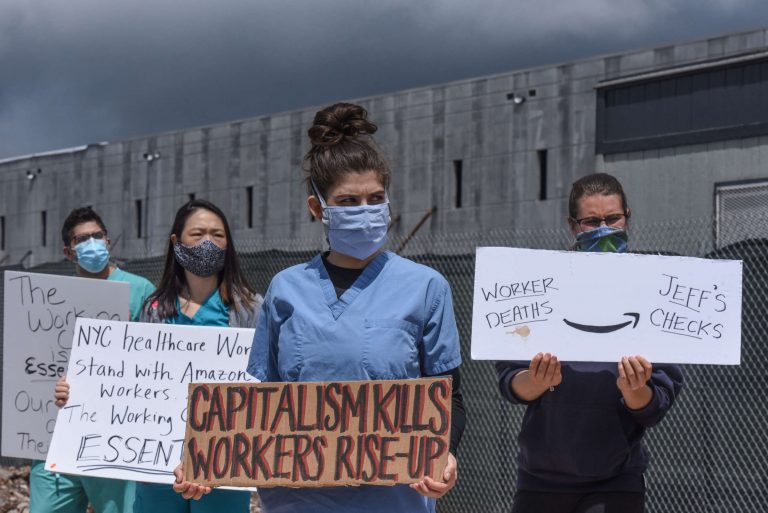 Four healthcare workers, including co-author of this article Mike Pappas, stand in scrubs and masks in front of an Amazon warehouse holding signs in support of better wages and safer working conditions for Amazon workers.