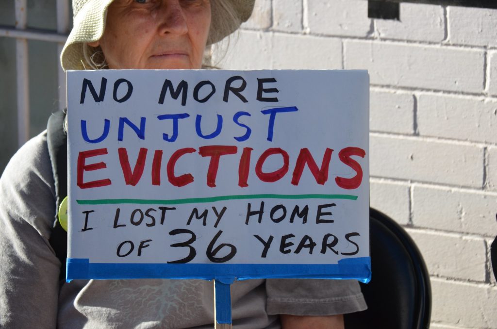 An older woman wearing a hat holds a sign that reads "No More Unjust Revictions: I Lost My Home of 36 Years"