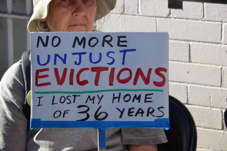 An older woman wearing a hat holds a sign that reads "No More Unjust Revictions: I Lost My Home of 36 Years"