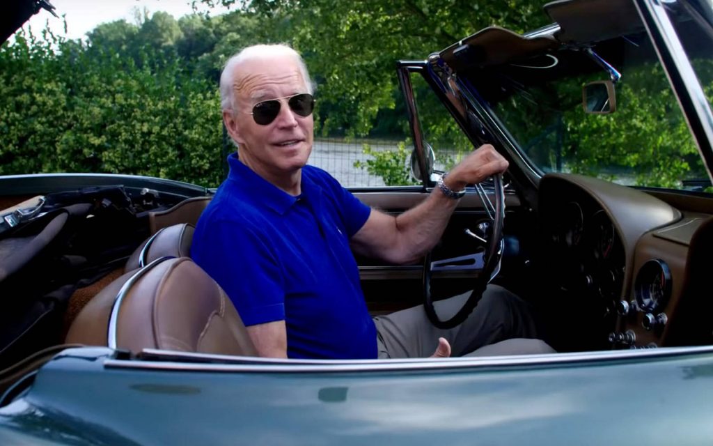 U.S. president Joe Biden sits in a sports car. He is wearing sunglasses and smiling.