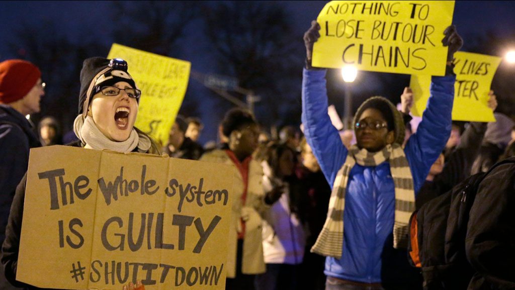 Protester holds a sign that says "The whole system is GUILTY #SHUTITDOWN. Another protester behind them wearing a blue sweater and yellow striped scarf holds a sign that reads "Nothing to lose but our chains."