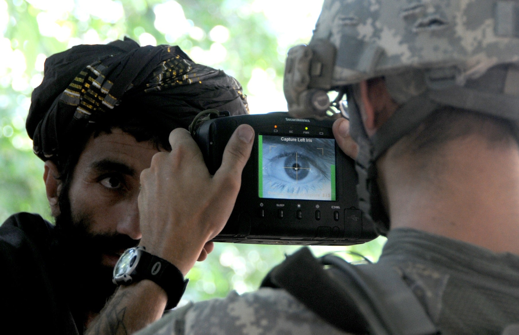 A U.S. soldier in army uniform and helmet is shown from behind while taking a close-up photo of the eye of an Afghan man with a bear and turban.