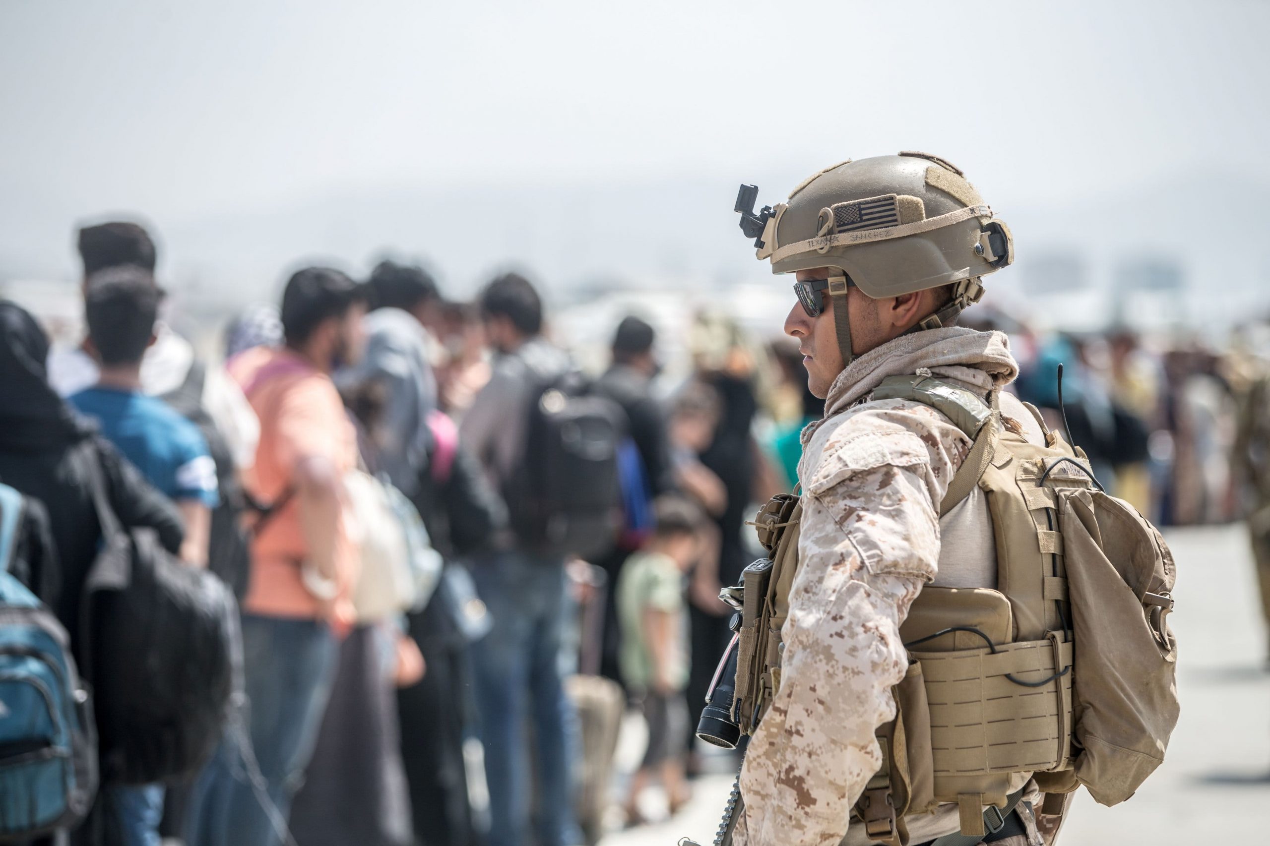 A U.S. soldier stands in the foreground in military gear and helmet. In the background, a line of Afghans.