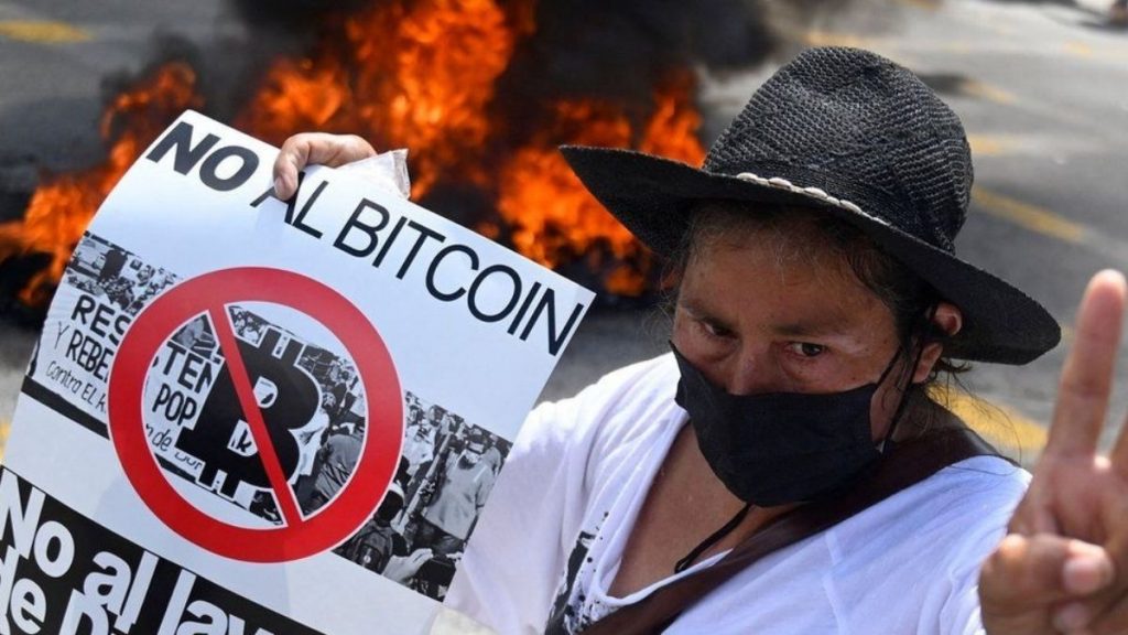 A woman in El Salvador wears a brimmed hat and a black mouth covering and holds a sign that says "No Al Bitcoin" (No to bitcoin). There is a fire behind her.