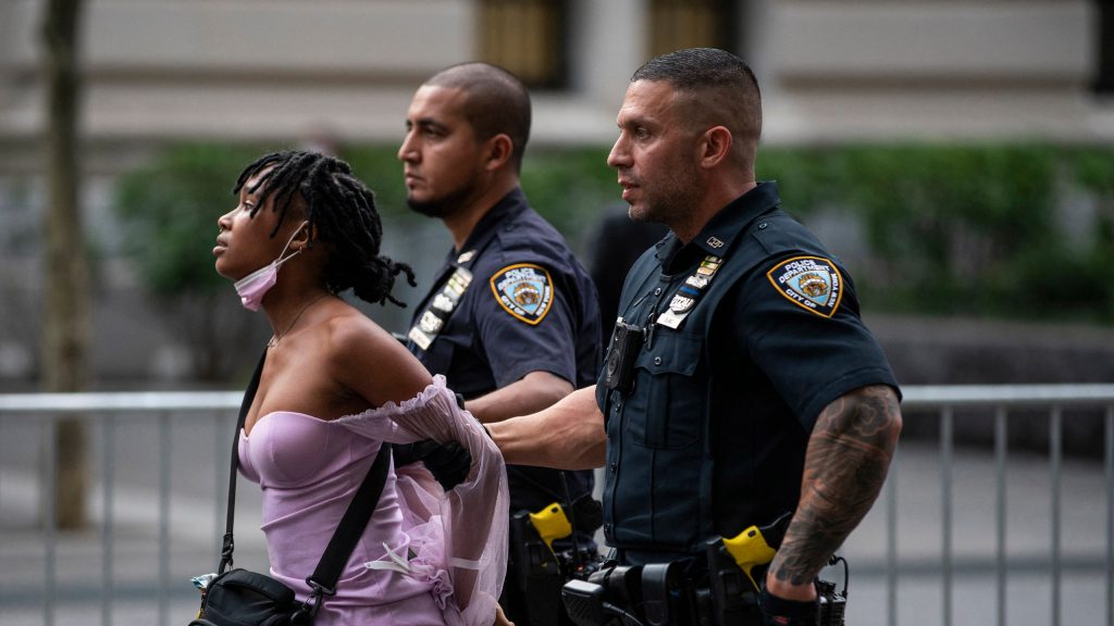 A black person wearing a purple shirt with a black crossbody bag is being restrained by two police officers.