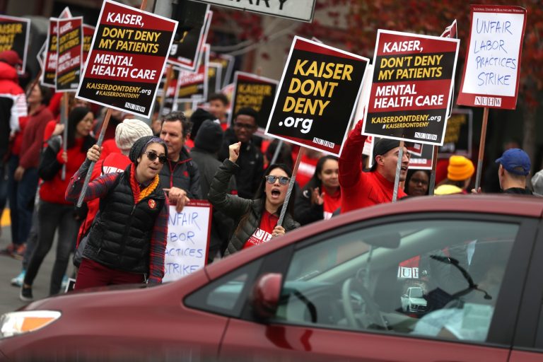 Healthcare workers dressed in red hold up protest signs against Kaiser Permanente while a car drives by.