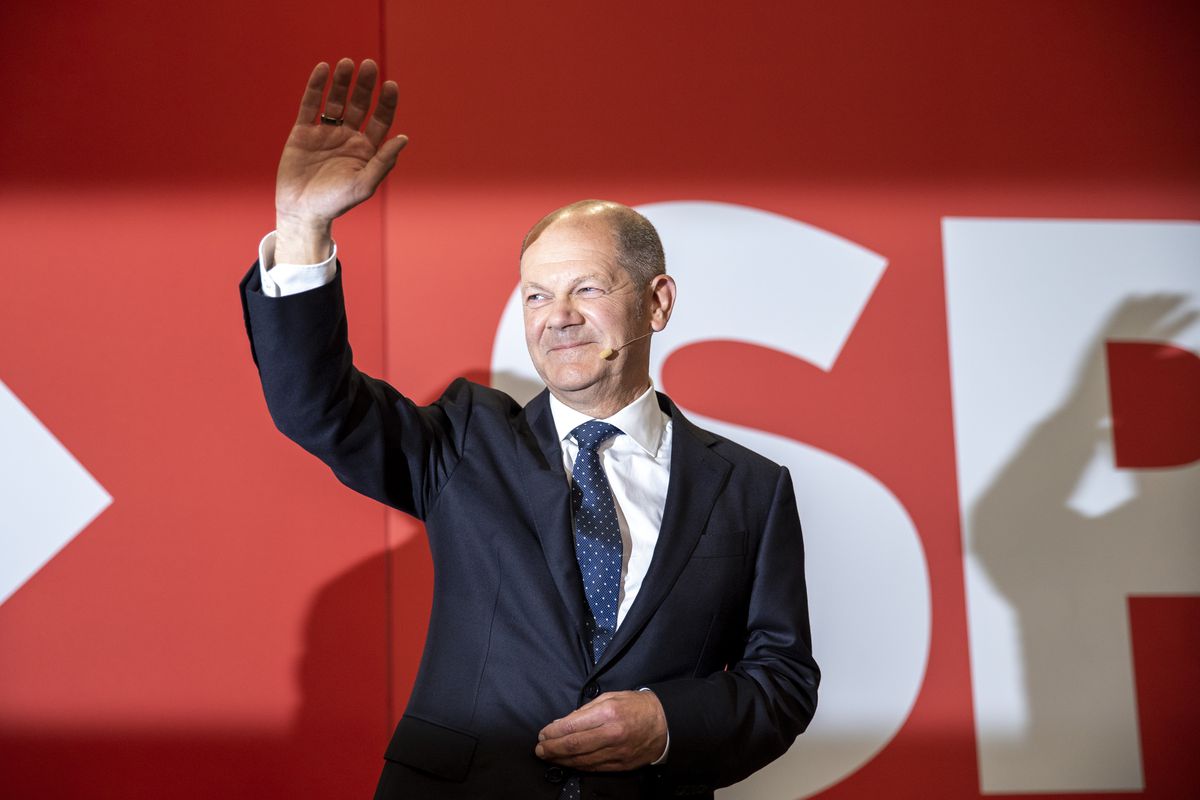 Standing in front of a red background with SPD written in white, Olaf Scholz, a balding man in his 50s, is wearing a suit, smiling, and waving at the audience.