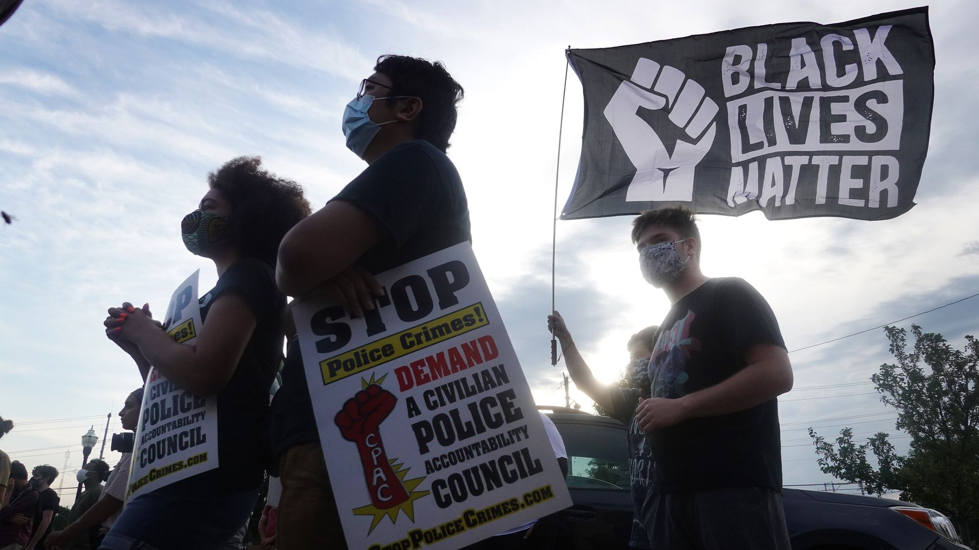 Four protesters of different races, all wearing face masks and black shirts, face to the left while holding Black Lives Matter signs. The sun shines brightly behind them in a partly cloudy sky