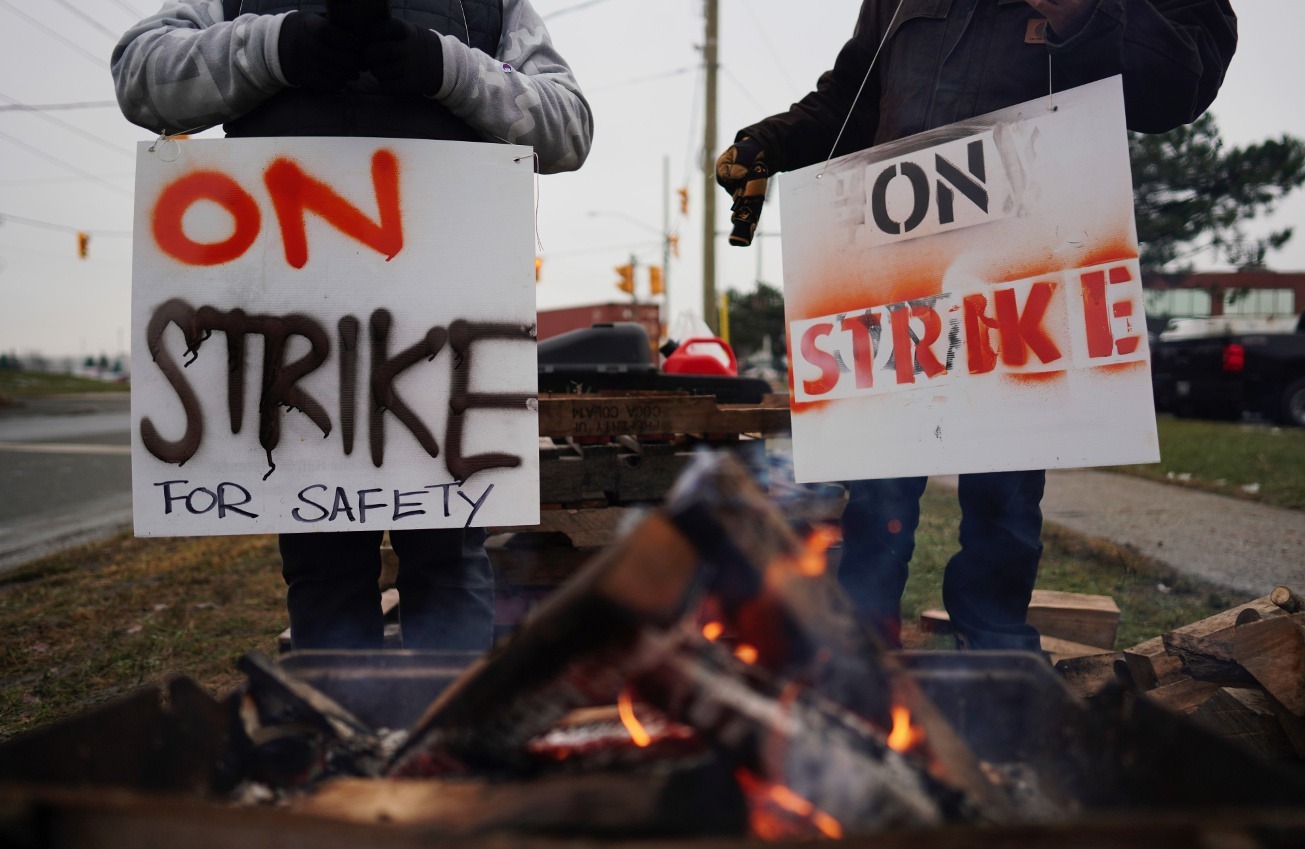 Two workers hold signs that say "On strike," in the foreground is a small fire.