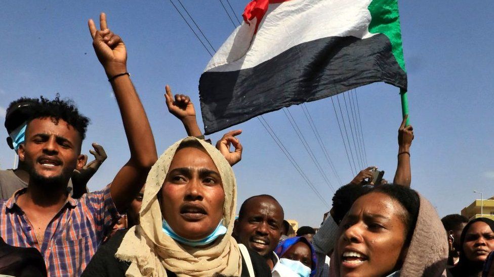 A Sudanese man and two Sudanese women marching against the coup in Sudan in October 2021. A Sudanese flag in the background.