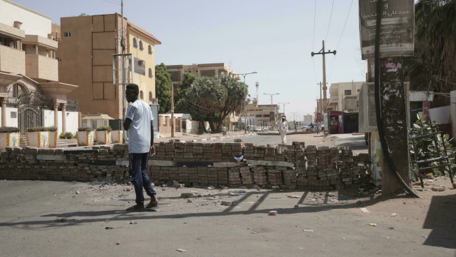 A lone Sudanese man stands on an empty street in daylight in front of a short wall of stacked bricks that spans the road