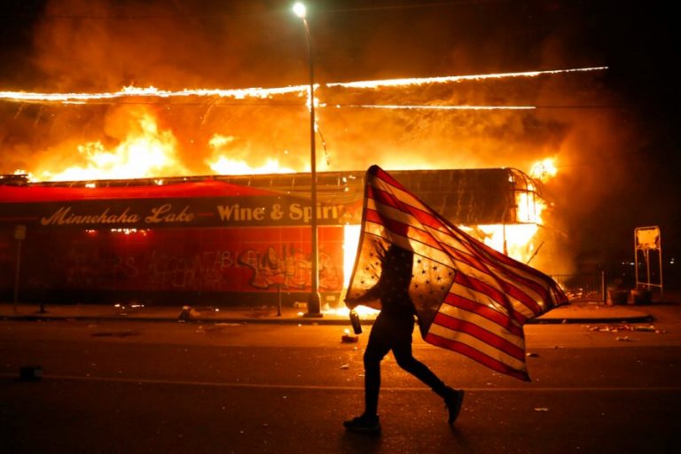 A burning building in Minnesota at night. A protester walks by with a flag.