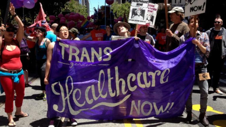 Trans rights protesters hold up a purple banner that says "Trans Healthcare Now"