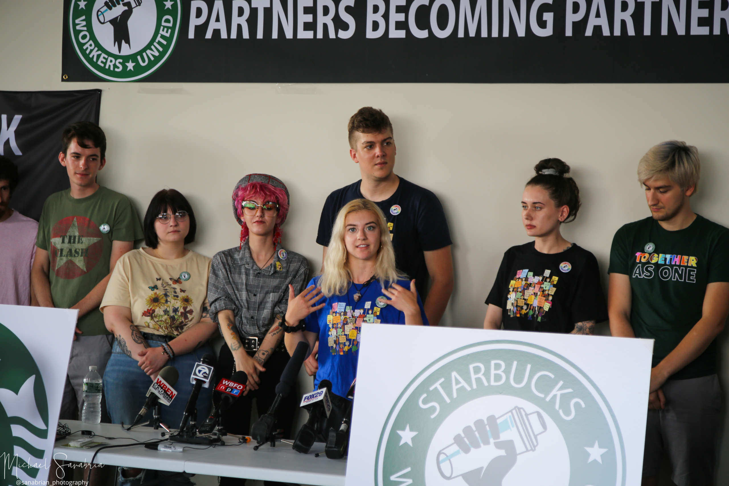Several Starbuck's employees speaking to reporters form behind a table.
