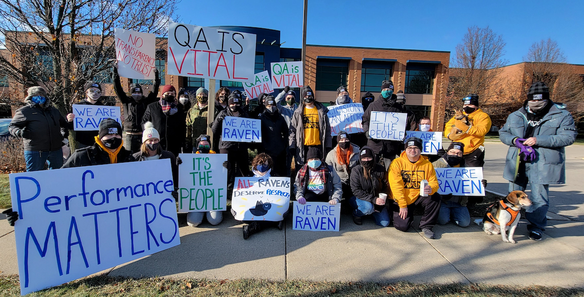 Group of about 20 workers dressed in winter clothing dressed with signs. Some signs read "RAVEN MATTERS", "QA IS VITAL", "PERFORMANCE MATTERS".
