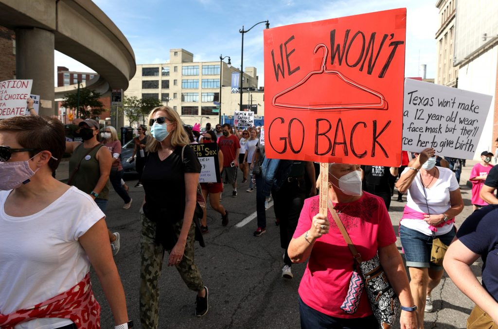 Group of protesters. A person wearing a white shirt with a red shirt tied around their waist is in the front left, and in the front right, a person wearing a red shirt is holding a red sign that reads "We won't" on top with a hanger in the center and "go back" on the bottom.