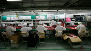 Workers dressed in white coats and round white hats sit at factory worktables on which there are red trays full of indistinguishable tech parts. The factory is dark except for the fluorescent lights over their tables.