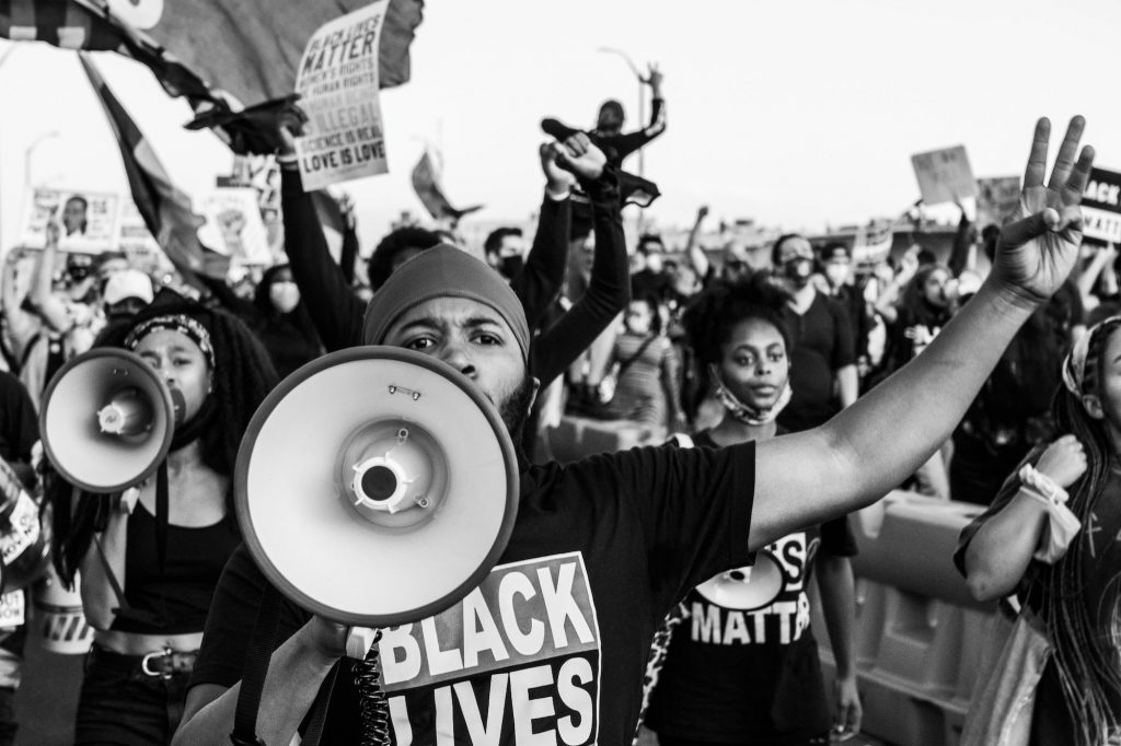 Black and white image, a Black Lives Matter protester leading a chant with a megaphone, people marching behind him.