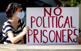 A white person wearing a mask, bandana, and sunglasses holds a white sign that says "NO POLITICAL PRISONERS" in large red letters