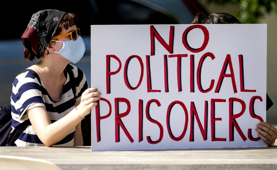 A white person wearing a mask, bandana, and sunglasses holds a white sign that says "NO POLITICAL PRISONERS" in large red letters